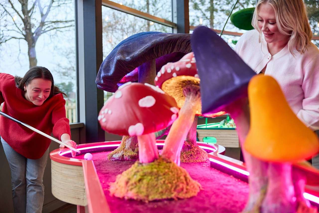 Two people play mini-golf, one aiming a putt while the other watches, around neon-lit lanes and oversized, colorful mushroom obstacles. Indoors near large windows, with trees outside and playful, glowing lighting.