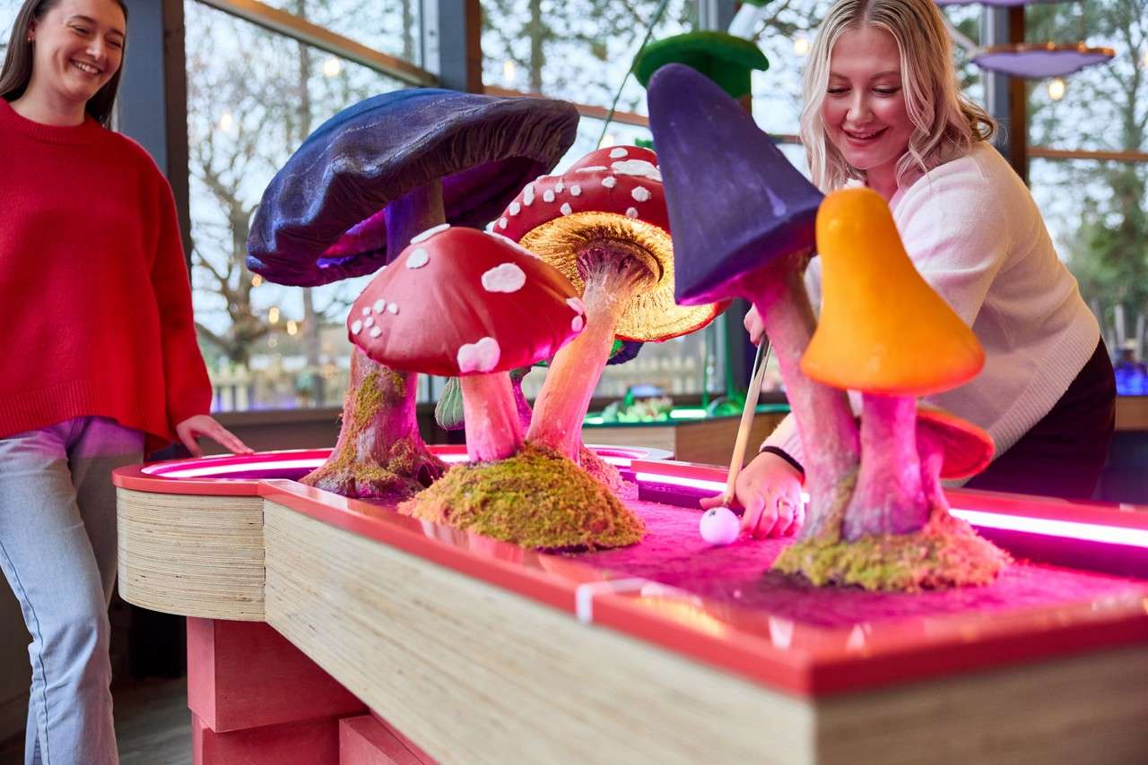 Two people putt a mini-golf ball through oversized, colorful mushroom obstacles, smiling and aiming around neon-lit edges, inside a whimsical indoor course with large windows and trees visible outside.