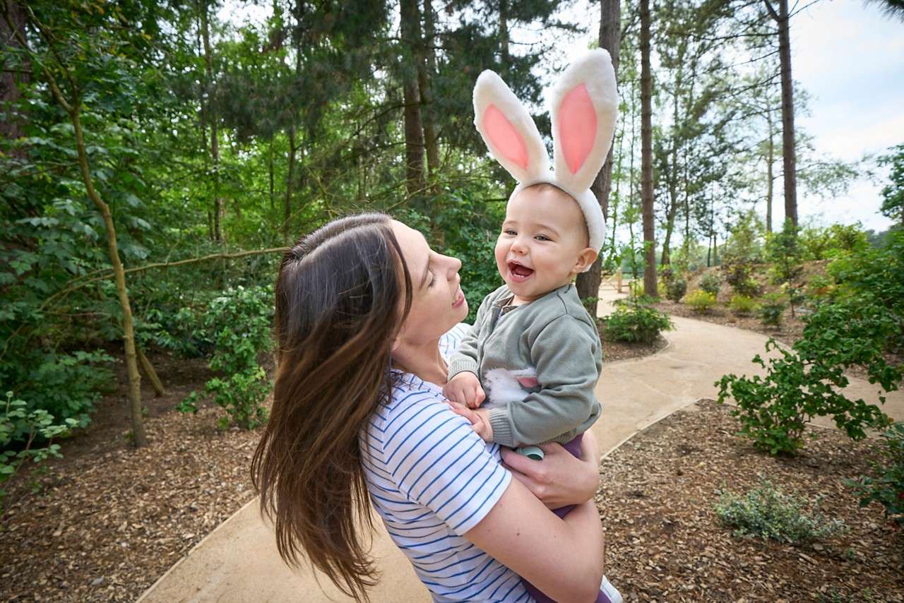 Smiling baby in bunny-ear headband laughs while being held by an adult on a winding path through a green forested park with tall trees, shrubs, and mulch.