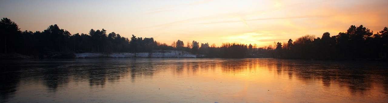 Lake reflects orange sunset; glassy surface mirrors the sky while dark, tree-lined shores frame the water in a calm, wide panorama at dusk.