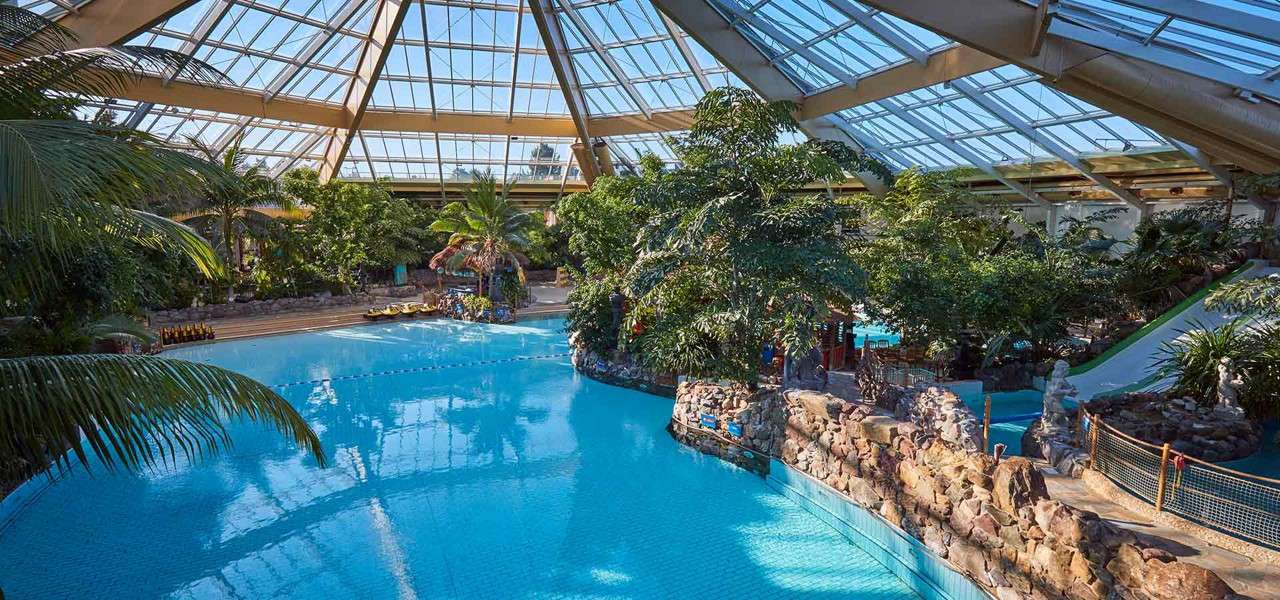 Indoor swimming pool glistens under a glass-domed atrium; water curves around rock-edged islands with tropical trees; nearby waterslide and deck chairs sit empty, creating a resort-like indoor oasis.