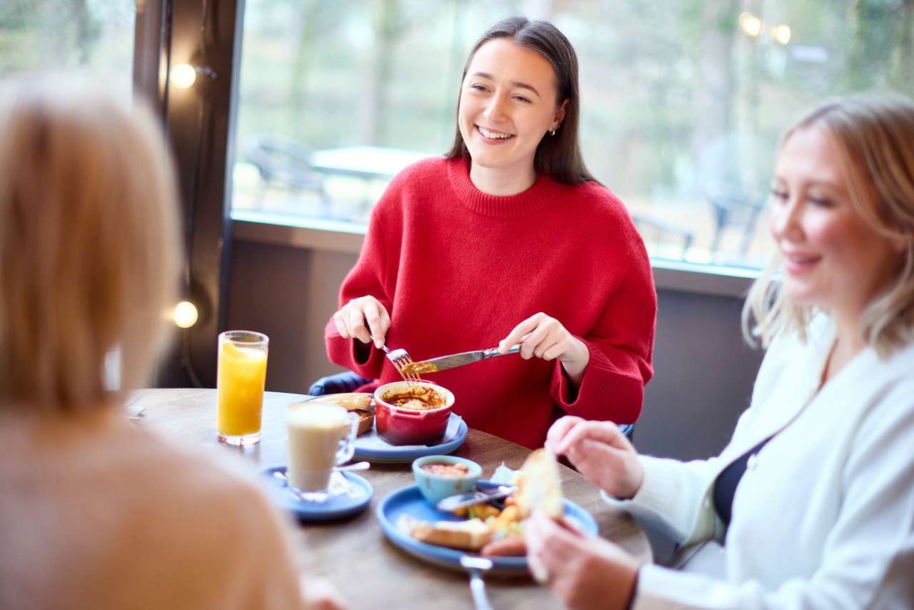 Smiling woman in red sweater eats with fork and knife. Two companions share breakfast at a cafe table with plates, coffee, and juice beside a large window with string lights.