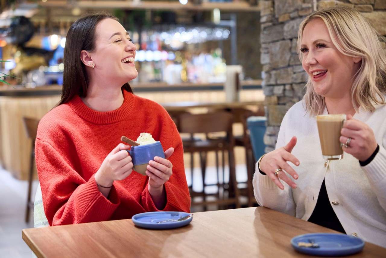 Two women laugh while holding hot drinks; one in a red sweater with a whipped-cream mug, the other with a latte, seated at a cafe table amid chairs and lights.