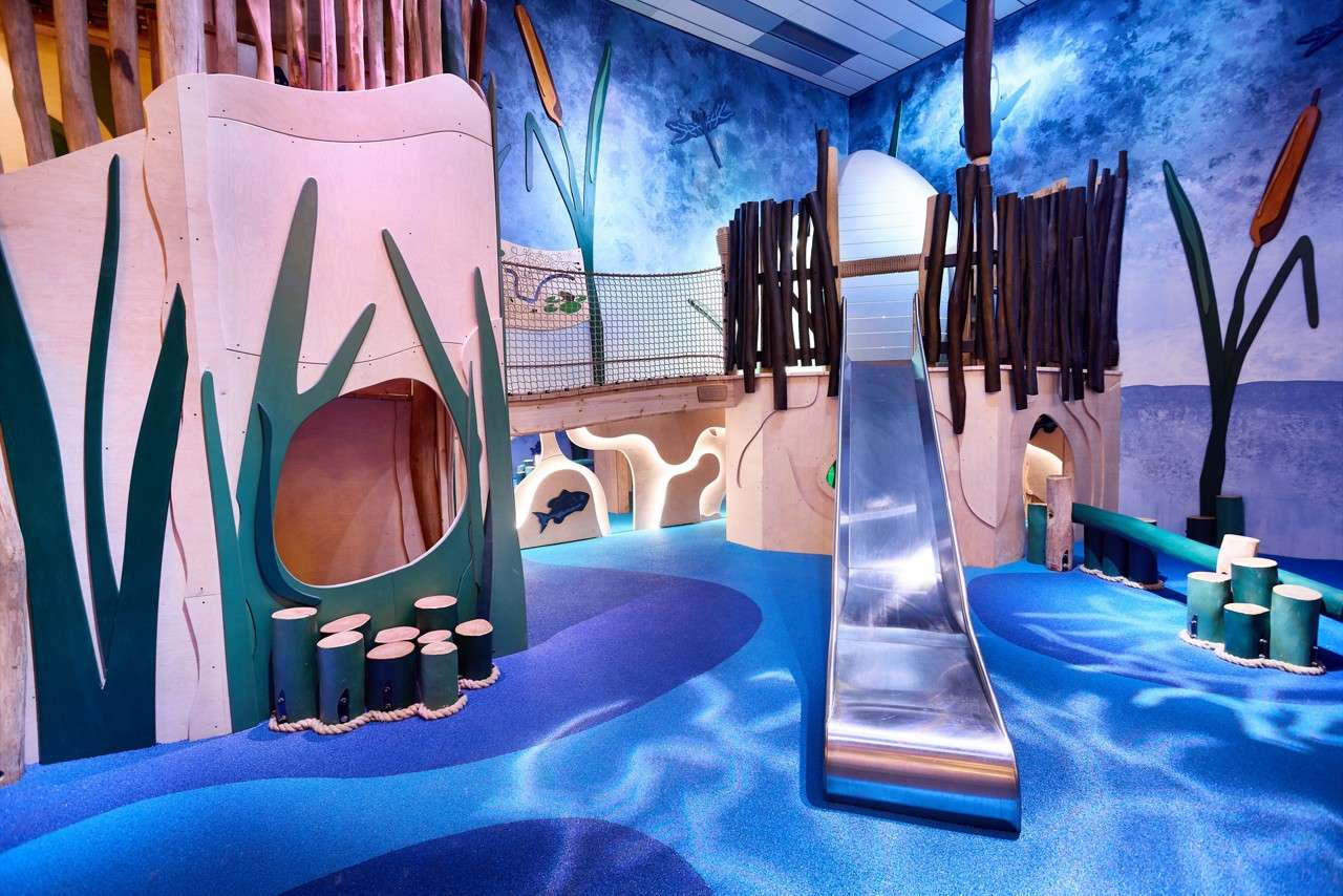 Metal slide descends from a wooden play fort, set within an indoor, aquatic-themed playground featuring blue wavy flooring, rope bridge, tunnel openings, cattail murals, and stump stepping posts.