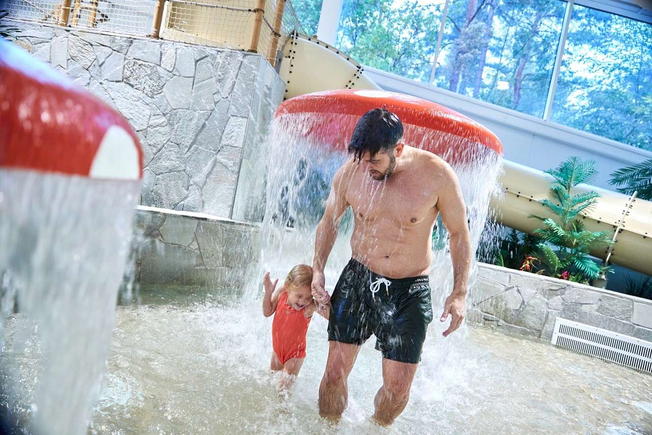 Man and child hold hands, stepping through a curtain of water from a red mushroom fountain; shallow indoor pool surrounds them with stone walls, waterslide tube, plants, and large windows.