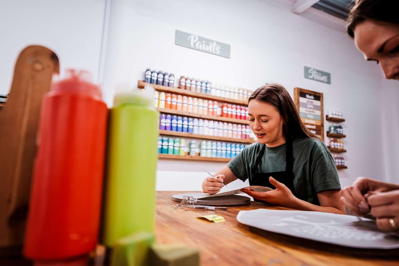 A person carefully cuts a stencil with a craft knife on a round board, in a studio lined with colorful paint bottles; signs read: Paints and Stains; squeeze bottles sit in the foreground.