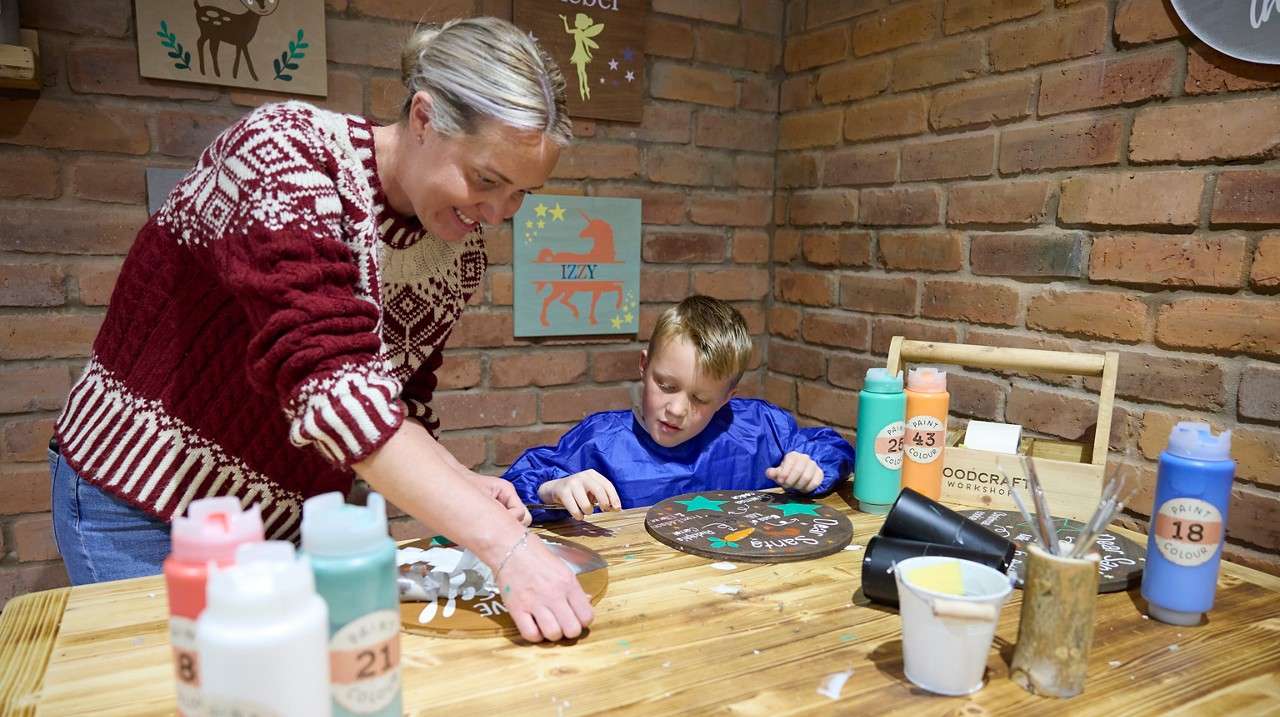 Adult and child paint wooden signs on a craft table in a brick-walled workshop. Text: IZZY; WOODCRAFT WORKSHOP; PAINT COLOUR 24; PAINT COLOUR 43; PAINT COLOUR 18.