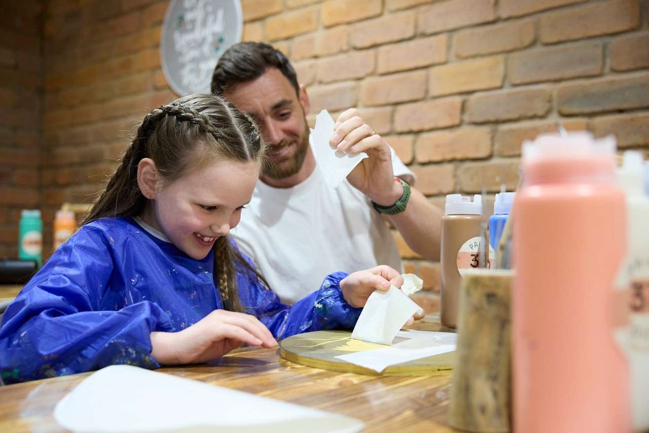 Woman aims a dart at a glowing interactive target in an arcade setting; screen text reads: UP NOW, ROUNDS, DAN, 6, 5, 6, WORTH, YE.