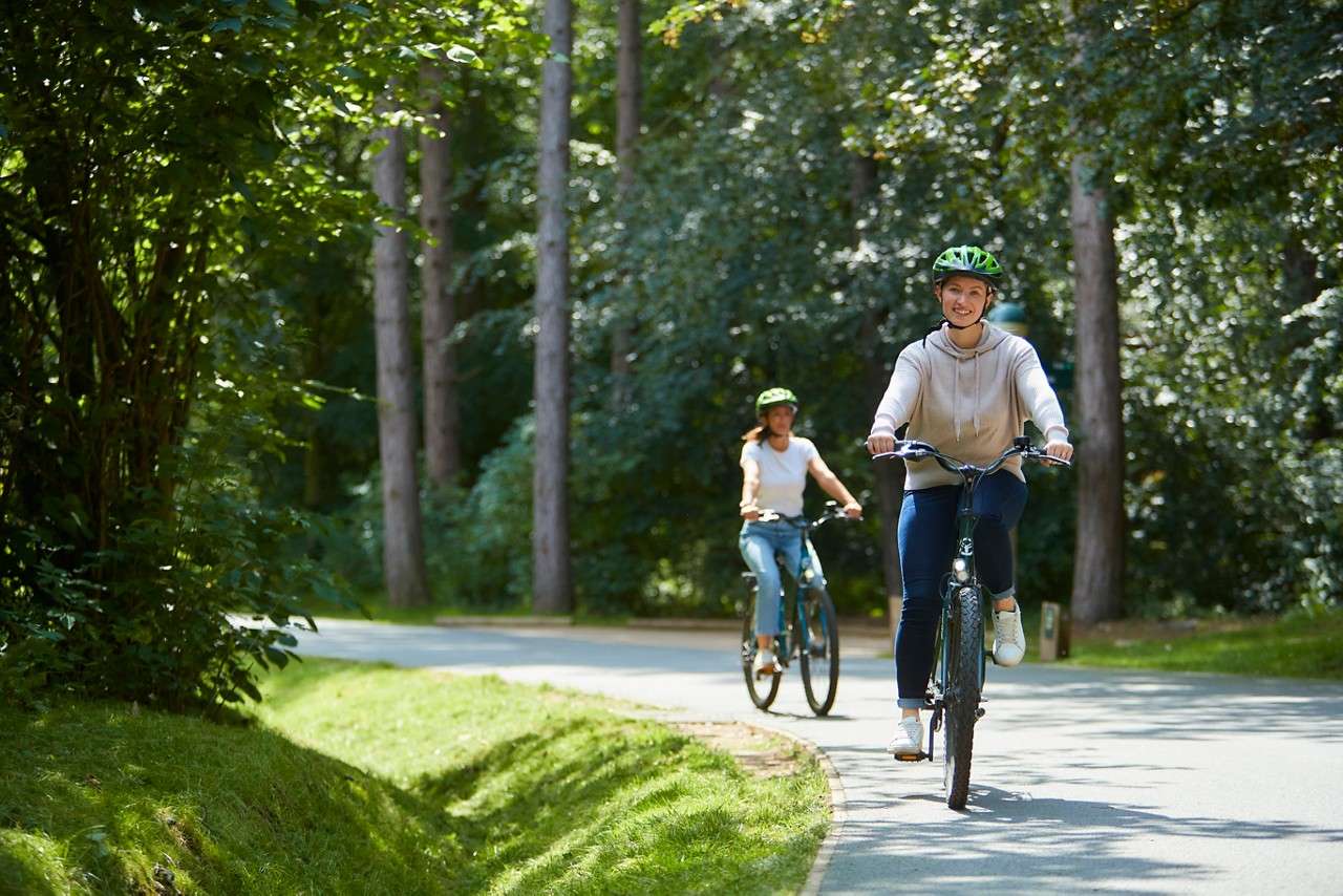 Two cyclists ride along a winding paved path through a sunlit forest, wearing helmets; the front rider smiles, trees and grass line the route, dappled light filtering through leaves.