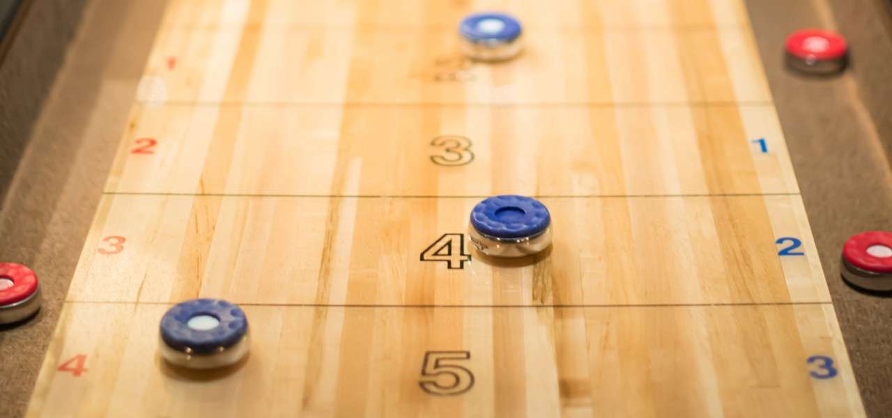 Shuffleboard pucks rest and slide on a varnished wooden board with scoring lines. Visible text: 3, 4, 5 (center); 2, 3, 4 (left); 1, 2, 3 (right).