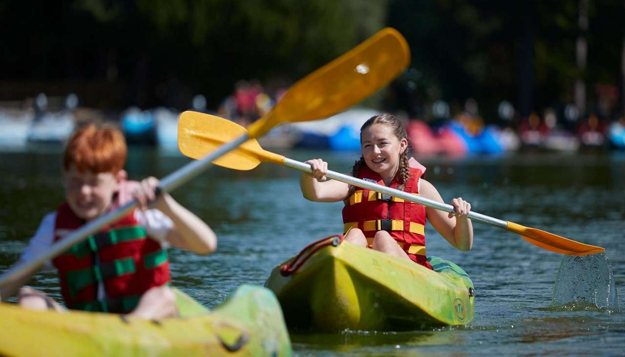 Children in kayaks paddle across a calm lake, wearing red life jackets; water splashes from paddles; blurred shoreline and colorful boats in the background on a sunny day.