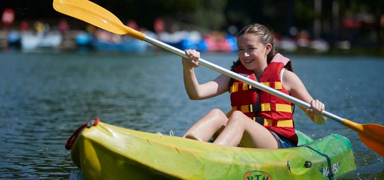 Kayaker paddles forward, smiling, in a green sit-on-top kayak on a calm lake; wearing red life jacket; background boats blurred. Visible text: RTM, MAMBO.
