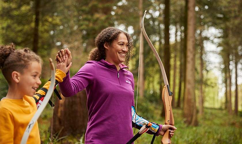 Archer holding a recurve bow high-fives a child, both smiling. In a forest setting with tall trees, they wear protective armguards and casual clothes, suggesting outdoor archery practice.