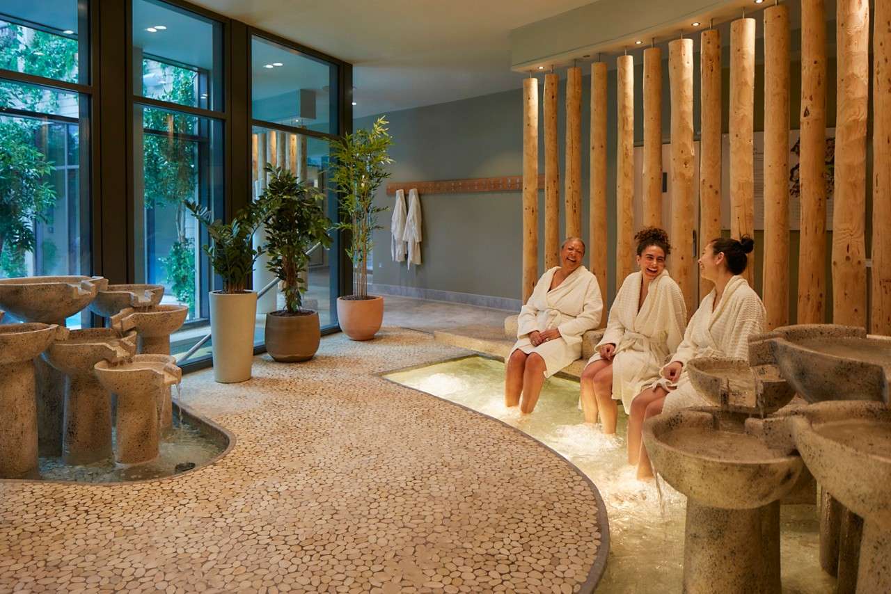 Three adults in white bathrobes soak feet and chat in a spa lounge; stone basins trickle water; potted plants, large windows, wooden columns, and a pebble floor create tranquil setting.
