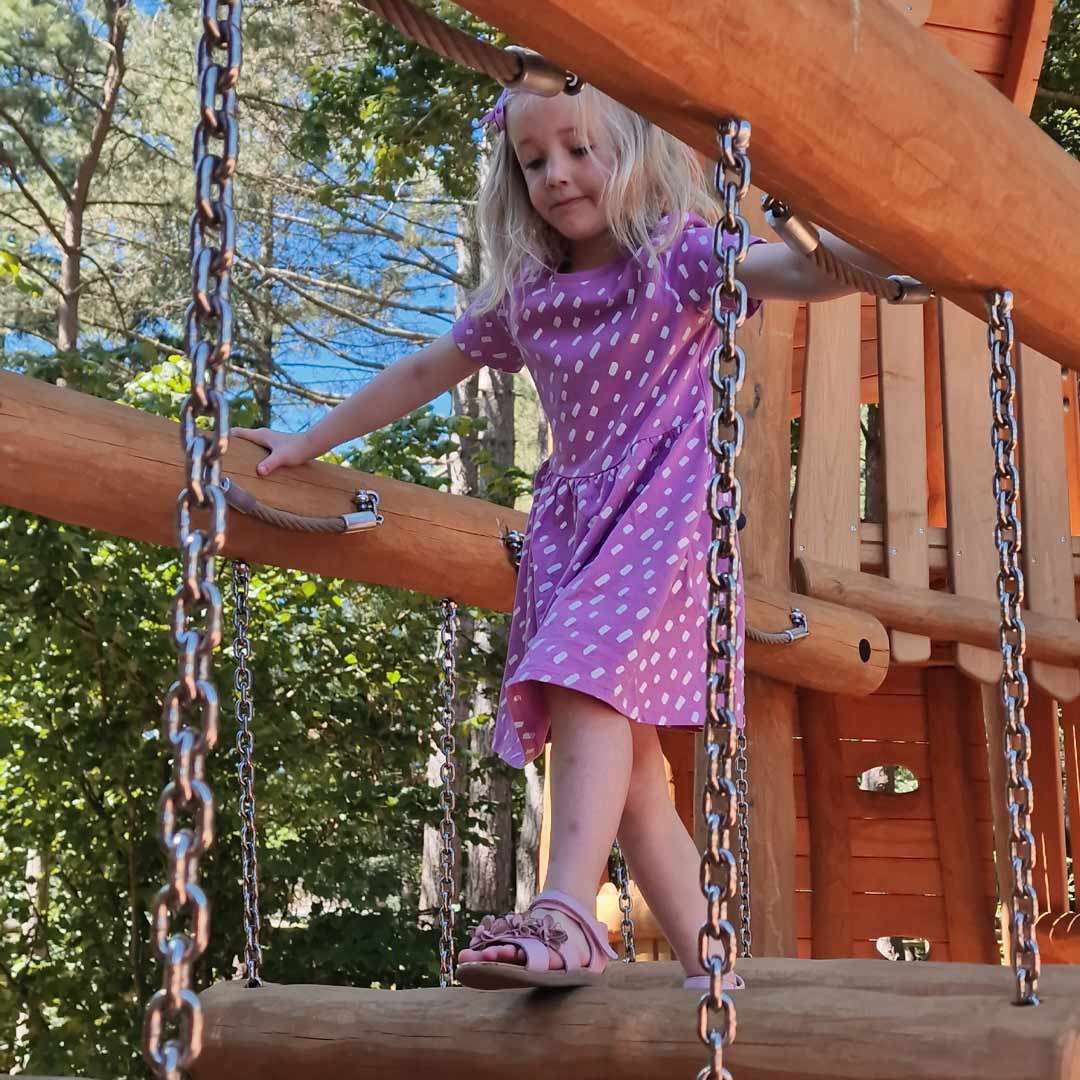Child in a purple dress balances on a chain-suspended wooden log bridge, holding side ropes; playground structure surrounds her, with sunlit trees and blue sky in the background.