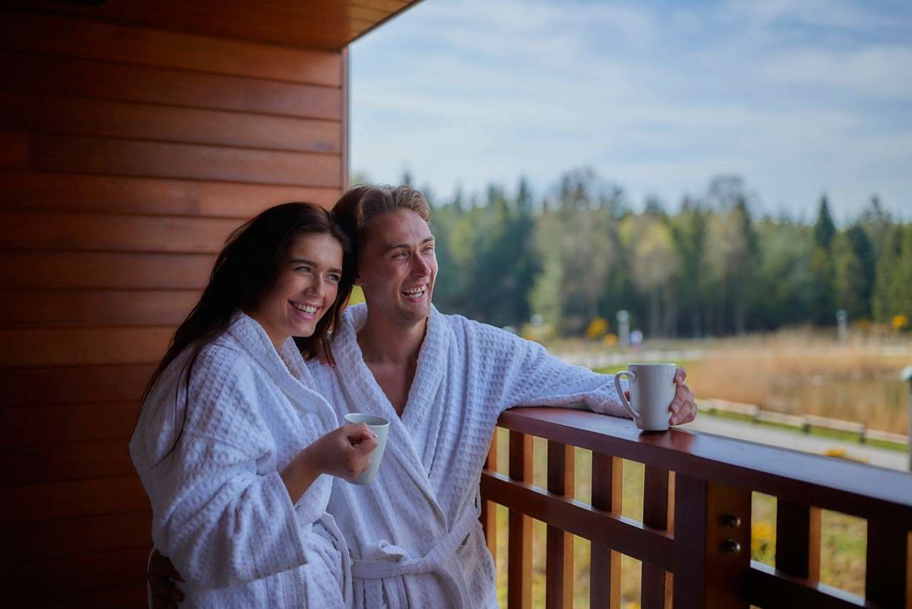 Two adults in white bathrobes smile and hold mugs, leaning on a wooden balcony railing, overlooking a forested landscape with meadow and road under a partly cloudy sky.