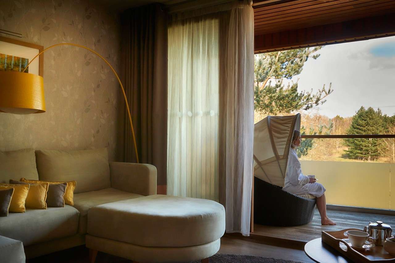 Person sits with a mug in a hooded wicker chair on a balcony overlooking trees; inside, a cozy room has a beige sectional, yellow lamp, sheer curtains, and teapot tray.