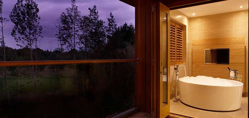 Oval freestanding bathtub filled with bubbles sits under warm lights, beside a handheld shower; context: wood-and-tile bathroom opens through sliding door to a glass-railed balcony overlooking trees at purple dusk.