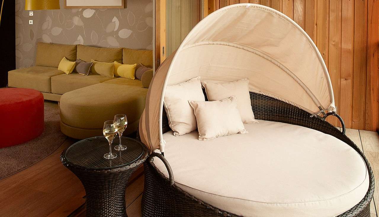 Round wicker daybed with beige canopy sits beside a woven table holding two glasses of white wine; behind, a tan sectional sofa, red ottoman, and wood-paneled wall form a lounge.