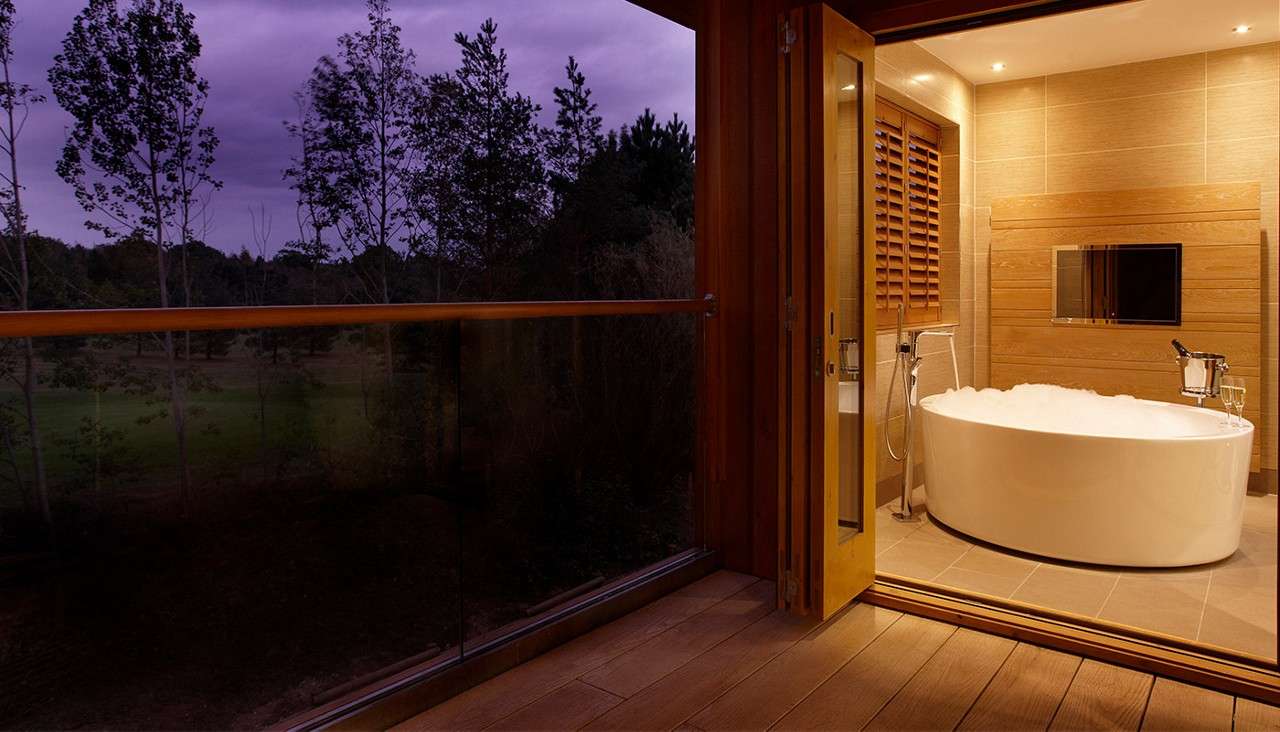 Bathtub filled with foam rests beside open sliding doors; a champagne flute on its rim, while wood-paneled bathroom with wall TV opens to a balcony overlooking trees at dusk.