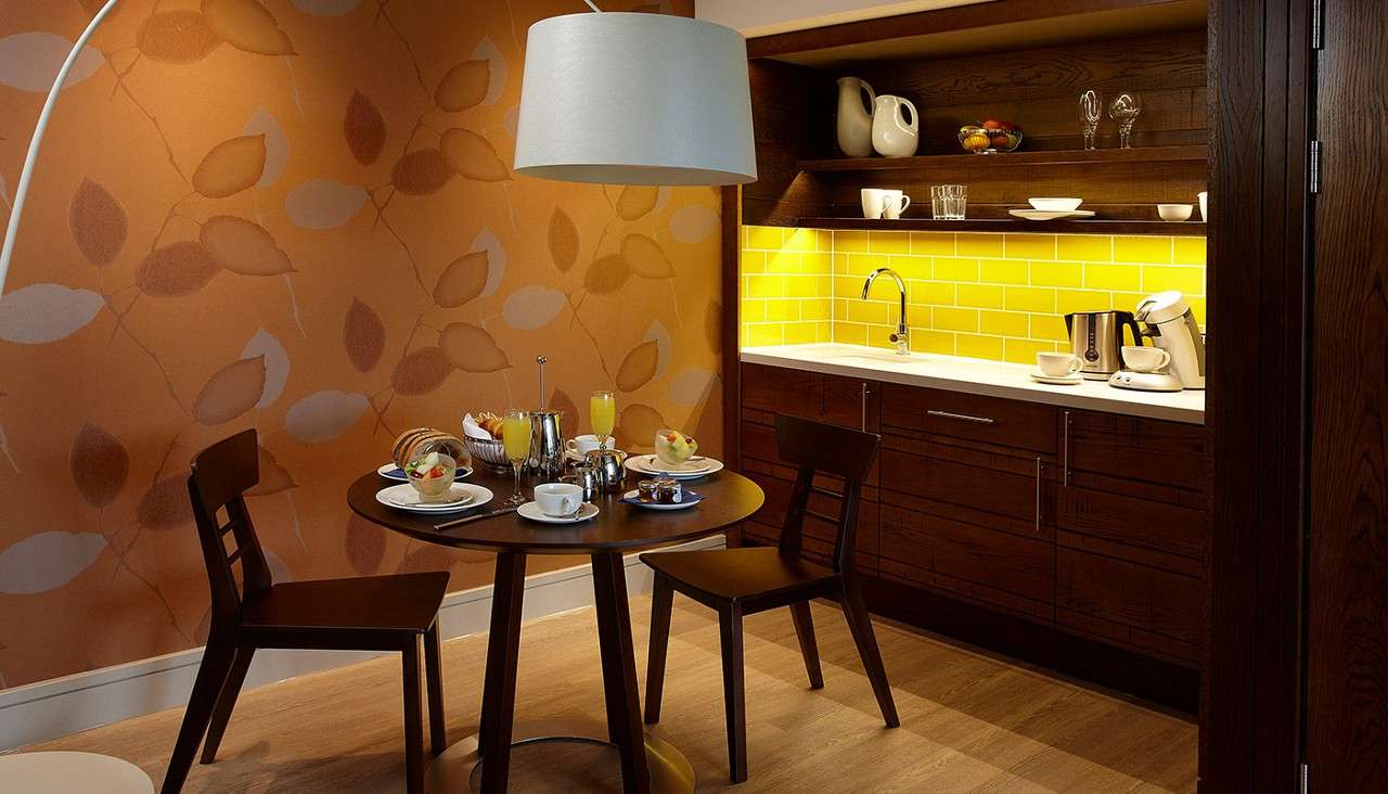 Round dining table hosts set breakfast—cups, fruit bowls, juice, teapot—beside a compact kitchenette with yellow-tiled backsplash, wooden cabinets, open shelves, and sink, under a pendant light, against leaf-patterned orange walls.