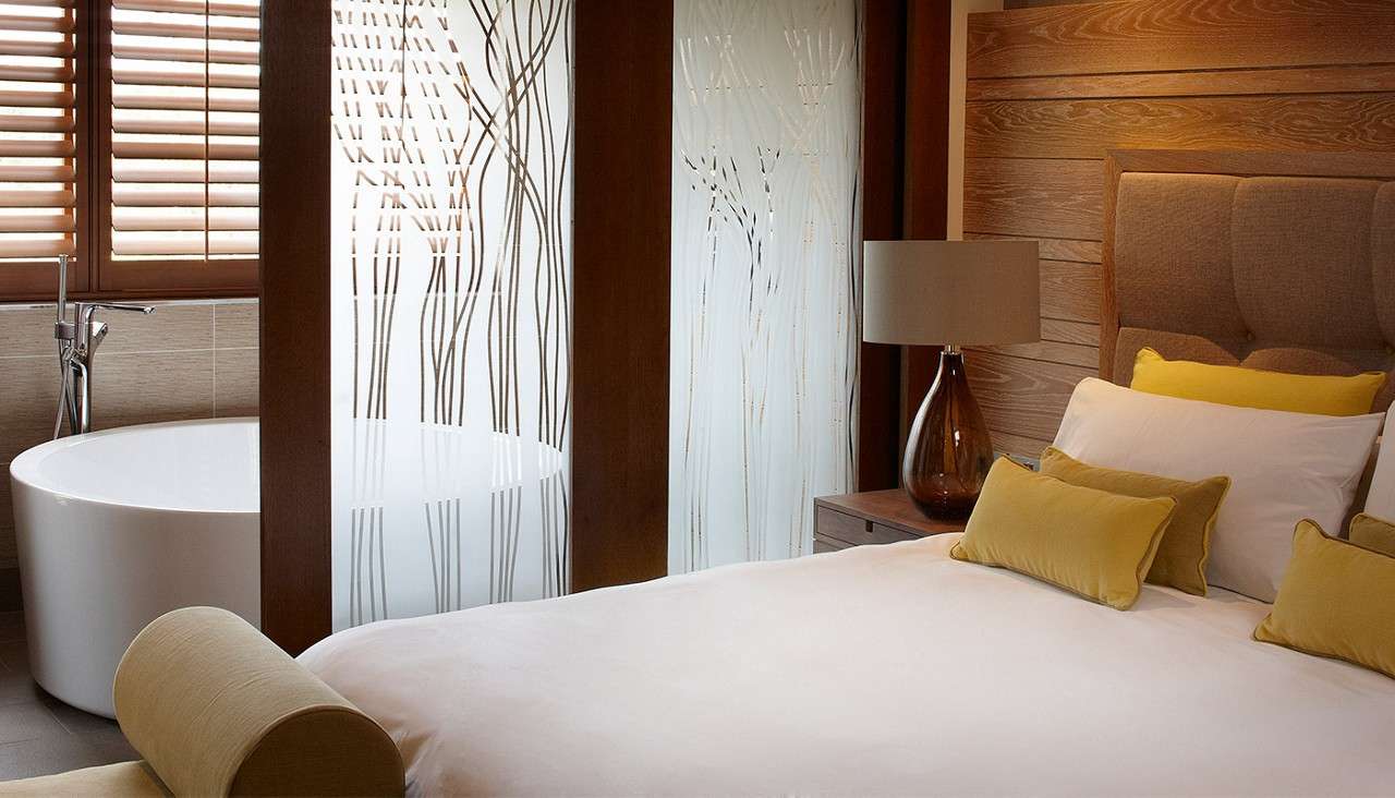 Bed with white linens and mustard pillows sits beside a bedside lamp; behind it, a freestanding bathtub partially obscured by frosted glass, with wooden shutters and warm-toned paneling surrounding.