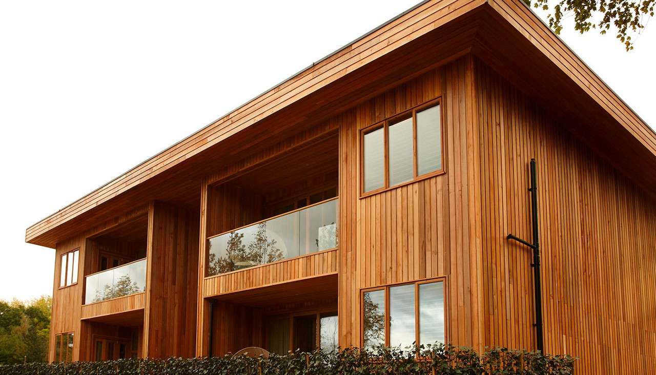 A modern two-story wooden house features vertical timber cladding and recessed balconies with glass railings, standing behind a hedge under an overcast sky, surrounded by trees.