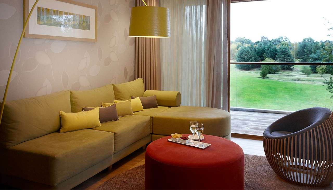 Sectional sofa anchors the space, supporting cushions and facing a red ottoman with two wine glasses; in a modern lounge with pendant lamp, leaf wallpaper, and window overlooking green parkland.
