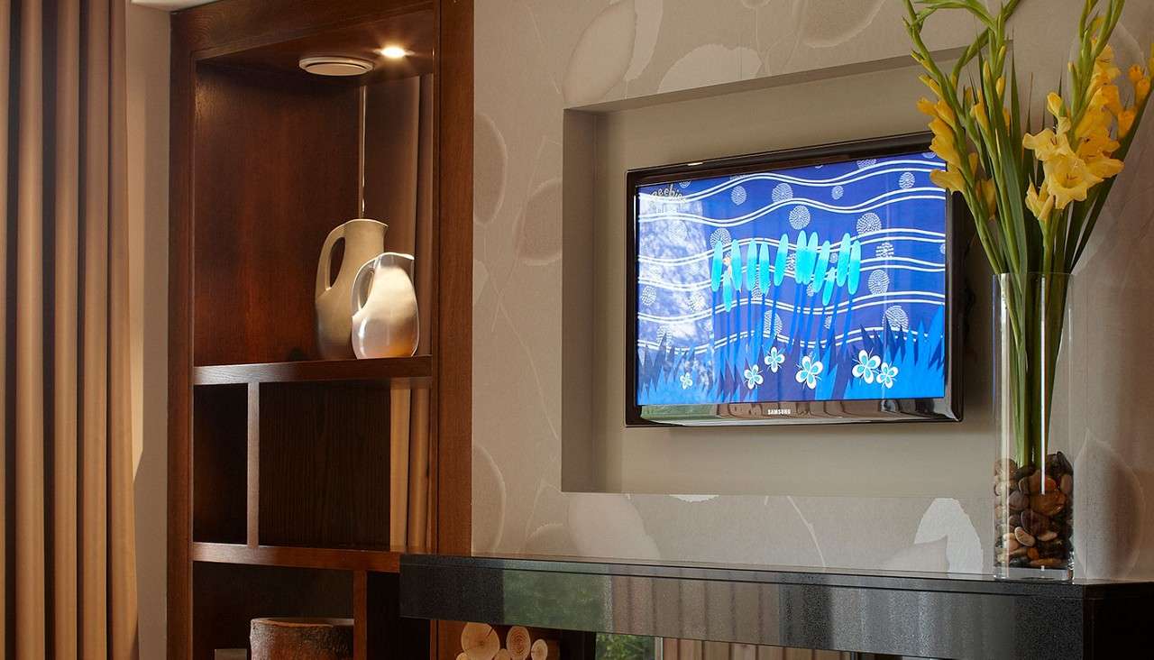 Wall-mounted TV displays blue floral animation; Text: SAMSUNG. In a living room with wooden shelving, ceramic vases, a glossy mantel, patterned wallpaper, and yellow flowers in a glass vase.