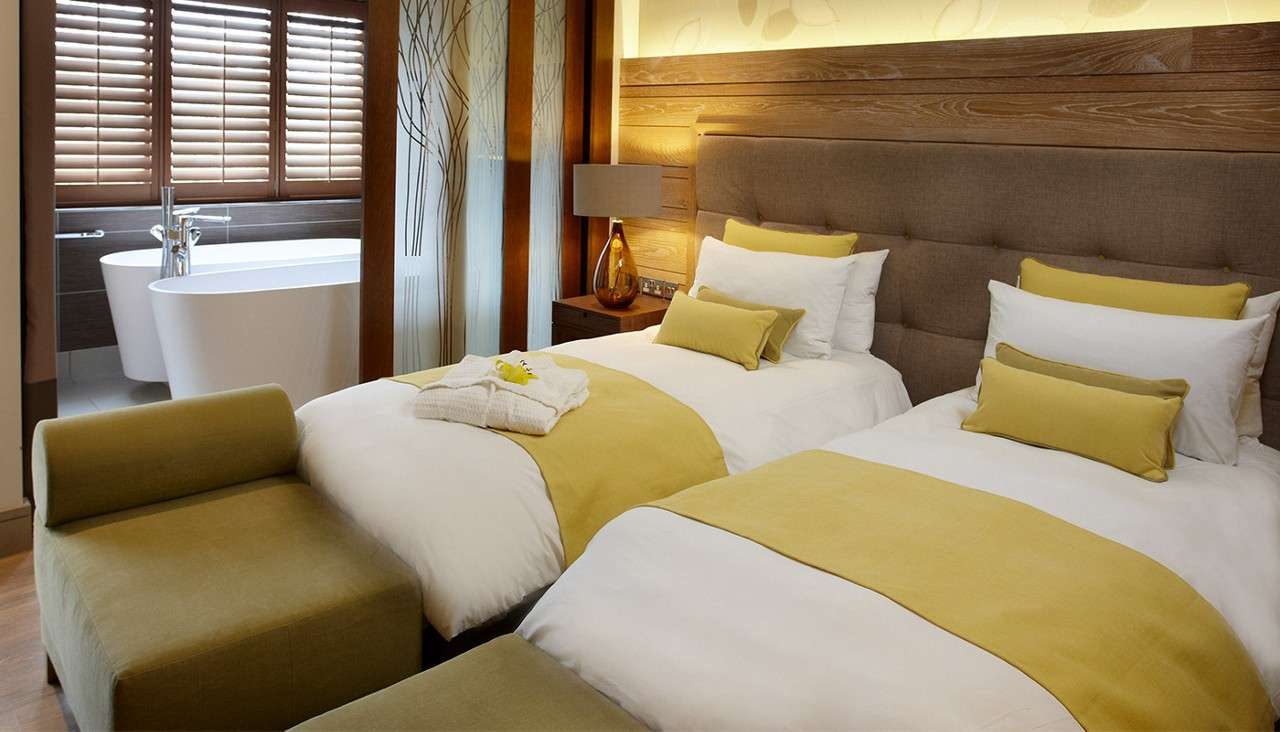 Two twin beds sit side by side, dressed in white linens with mustard throws and pillows; a bench faces them; through a wooden partition, a bathtub stands by shuttered windows.