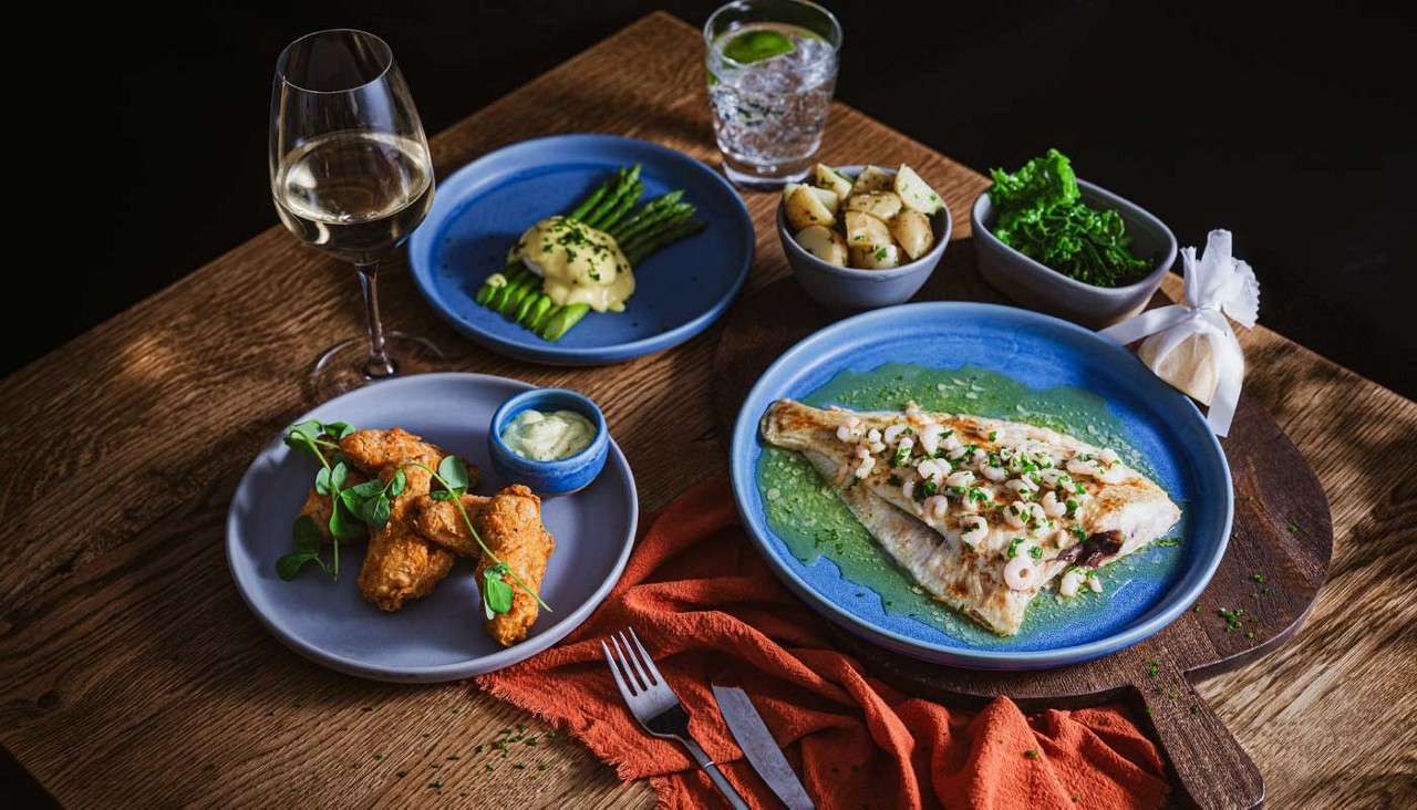 Grilled white fish topped with shrimp rests in green sauce, accompanied by asparagus with hollandaise, new potatoes, greens, and fried bites with dip; wine and cocktail on a wooden table.