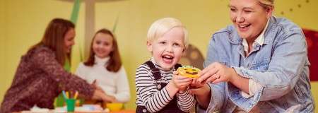 Child holds a decorated cookie and smiles while an adult assists, in a bright craft room with colorful walls; background people work at a table with cups and art supplies.