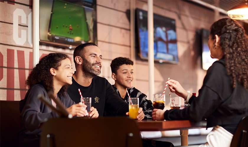 Four people sit at a table, smiling and chatting while drinking sodas with striped straws; background TVs show sports, and warm lighting and wood paneling suggest a casual sports bar.