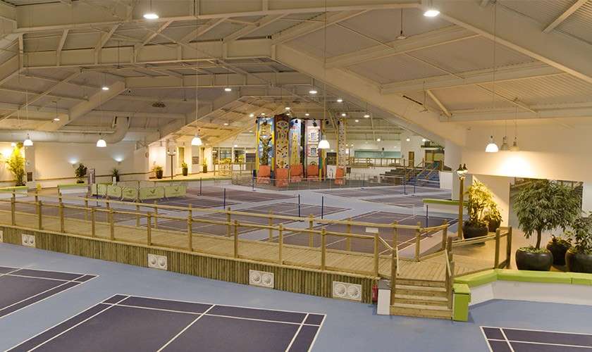 Multiple empty badminton courts sit under bright lights in a spacious indoor sports hall, bordered by a wooden viewing deck, potted plants, and a central colorful climbing structure.