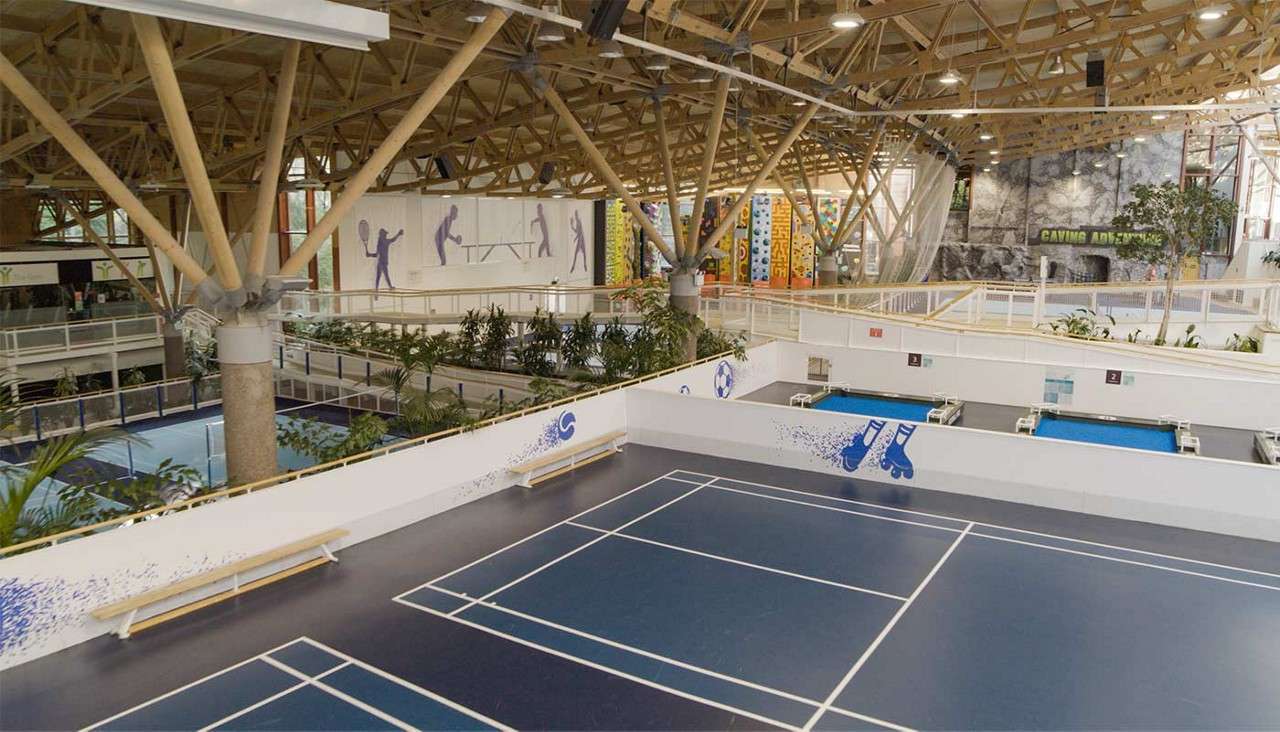 Badminton court sits empty, bordered by benches, while surrounding areas feature climbing wall, colorful climbing towers, plants, and walkways under a timber-truss ceiling; visible text: CAVING ADVENTURE, 2.