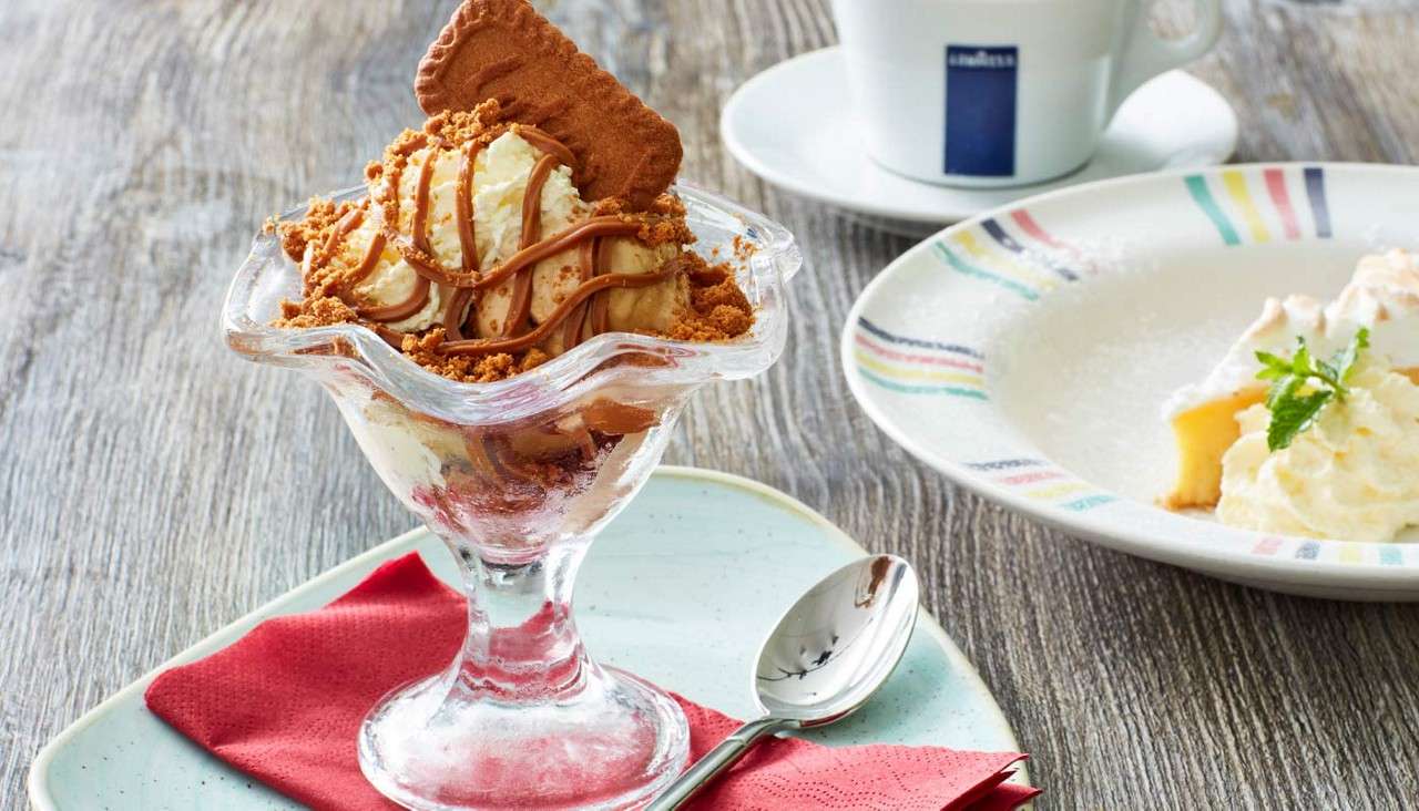 Sundae topped with caramel and crumbled biscuit sits in glass dish; spoon on plate with red napkin; background shows coffee cup and cake slice on wooden table.