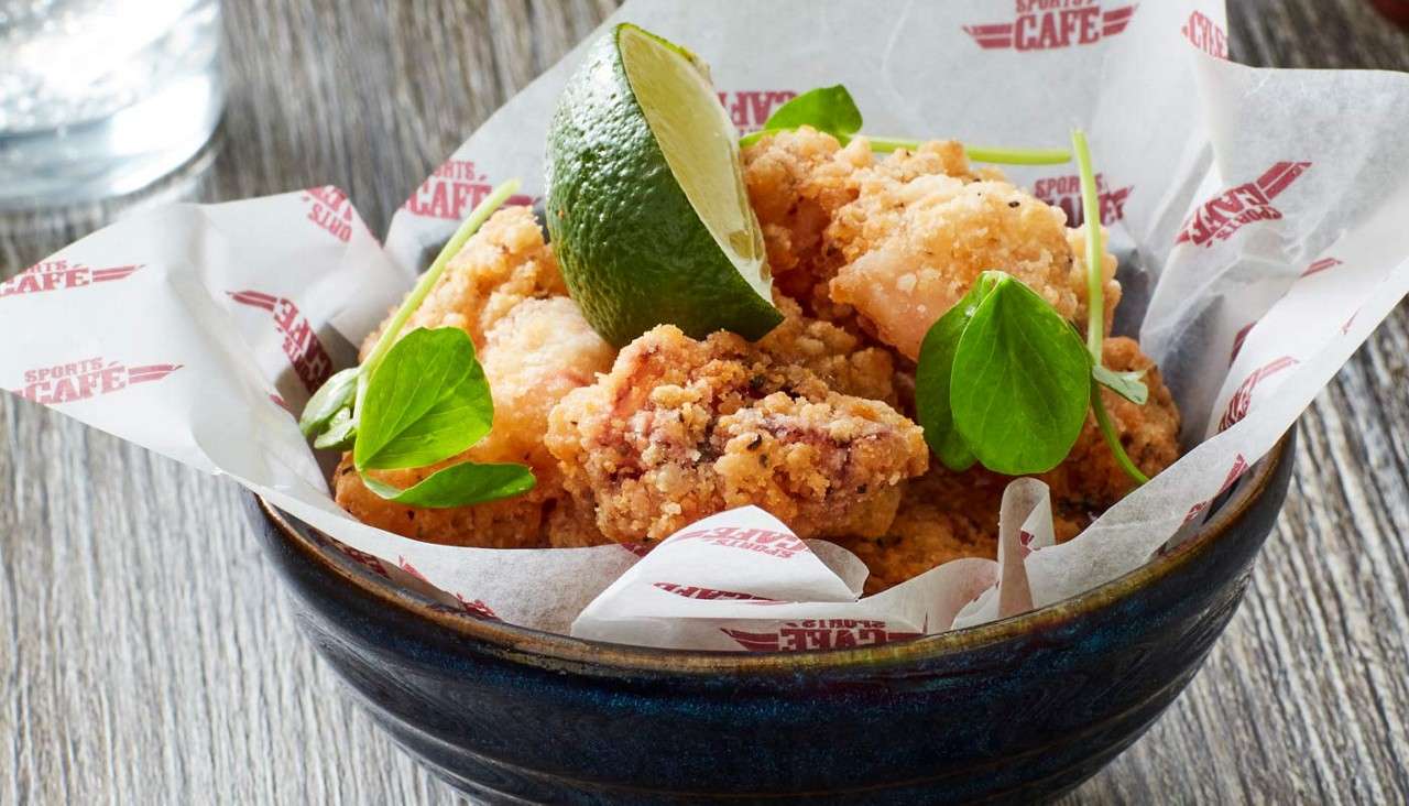 Crispy battered bites sit in a bowl, topped with a lime wedge and small greens, served on branded paper on a wooden table. Visible text: “SPORTS CAFE” (repeated).