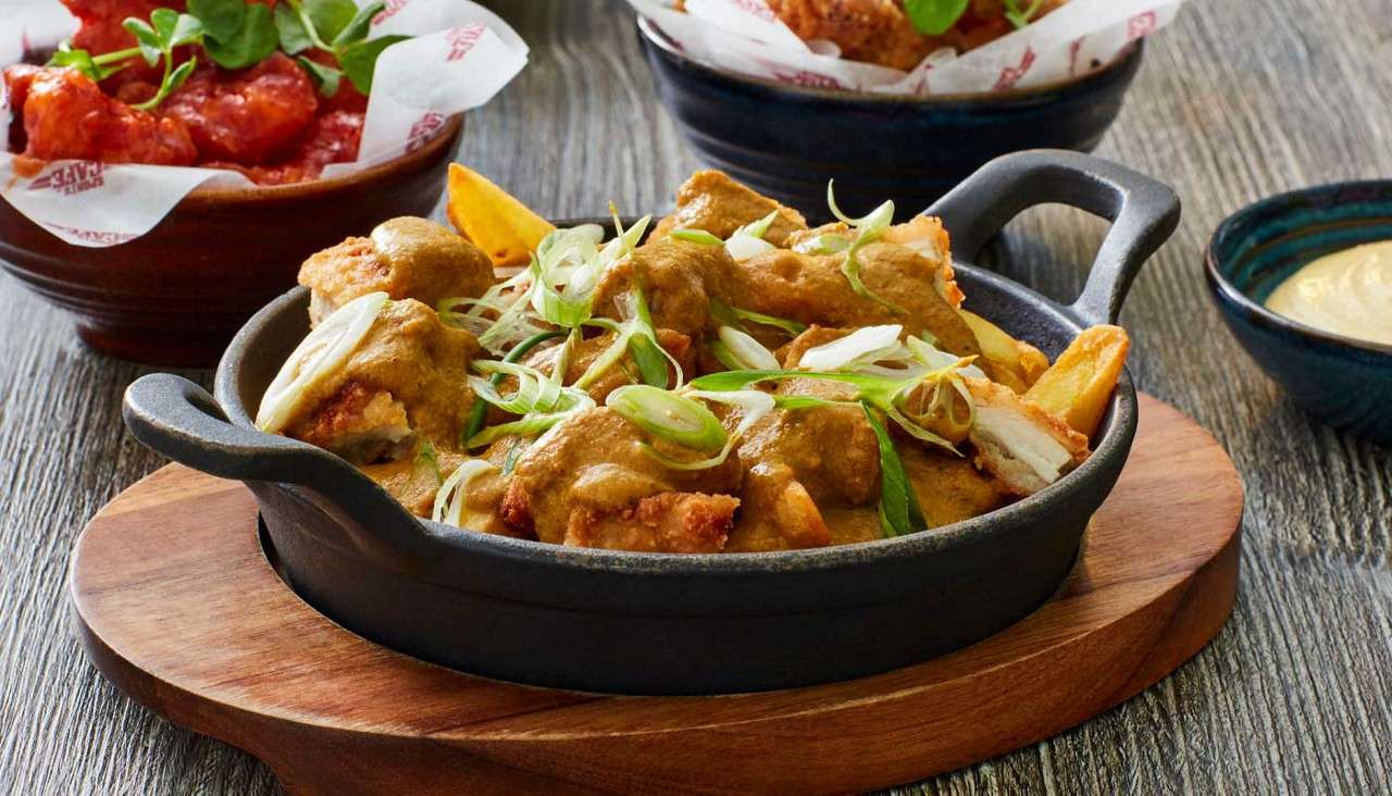 Crispy chicken pieces and fries are covered with curry sauce and topped with sliced scallions, served in a black skillet on a wooden board, with side dishes and dip.