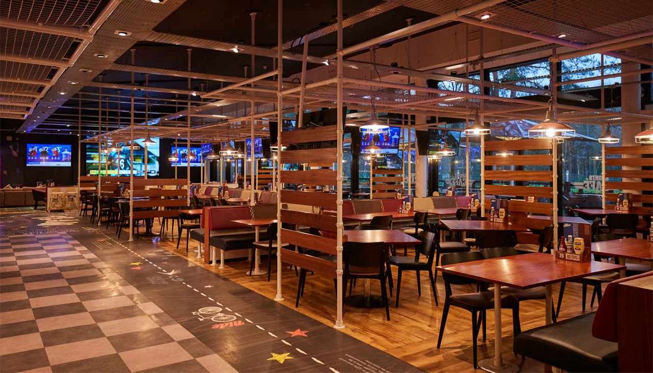 Tables and booths sit empty, lit by pendant lamps; a metal frame grid divides seating in a modern sports bar with many TVs and a patterned walkway. Text: 26; SPORTS BAR (reversed).