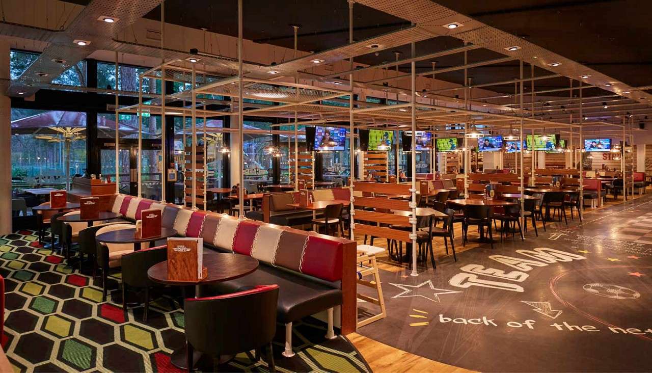 Sports bar seating sits empty, with wooden booths and round tables; many TVs show games. Chalk-style floor graphics read "TEAM" and "back of the net" in a spacious, window-lined interior.