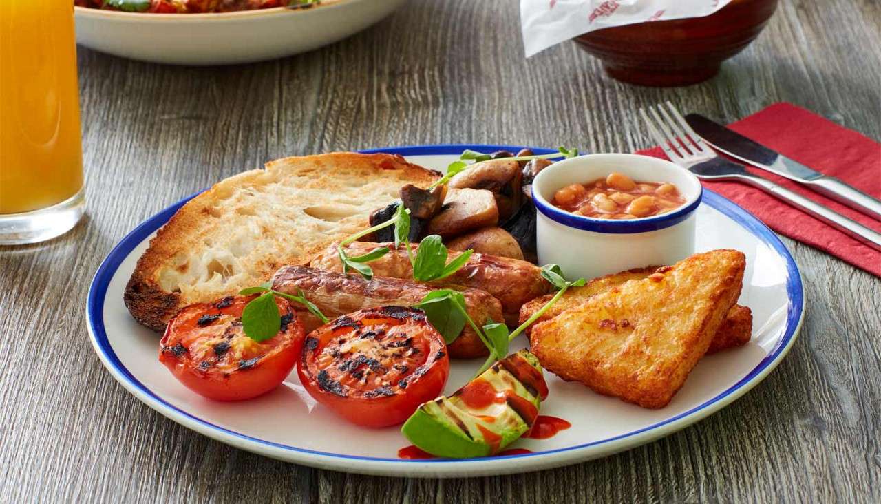 Breakfast plate presents sausages, grilled tomatoes, toast, mushrooms, hash browns, beans, and courgette with sauce, garnished with greens, served on a wooden table beside orange juice, cutlery, and napkin.