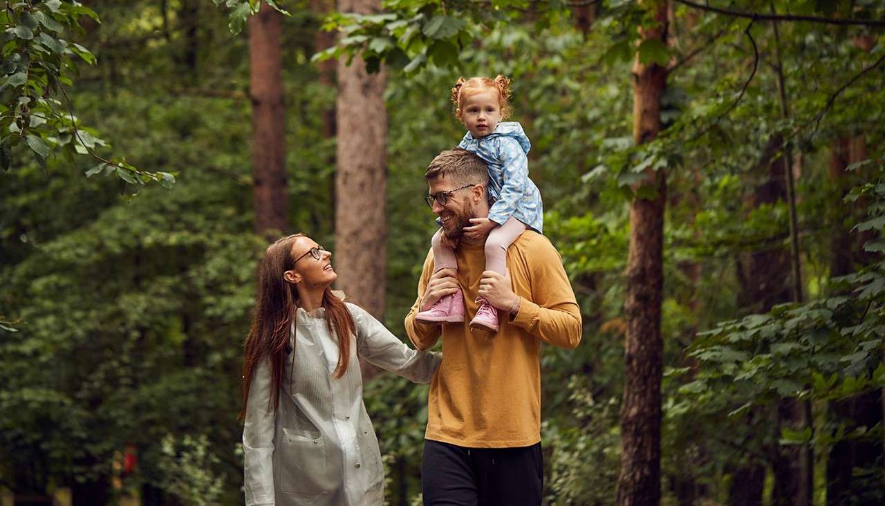Man carries a small child on his shoulders while a woman walks beside them, smiling. They stroll along a forest path surrounded by tall trees and dense green foliage.
