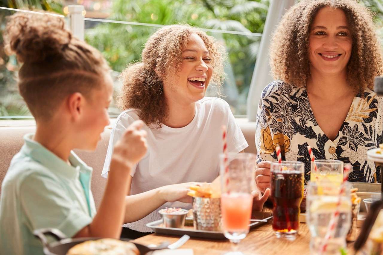 Three people sit at a restaurant table, laughing while eating fries and drinking sodas with striped straws. Sunlight and green foliage fill the background through windows, creating a relaxed setting.