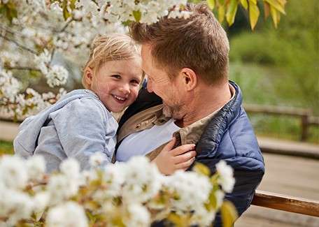 Adult man cradles a smiling child, both looking at each other affectionately, amid white blossoms in a park, with wooden railing and greenery in the soft background.