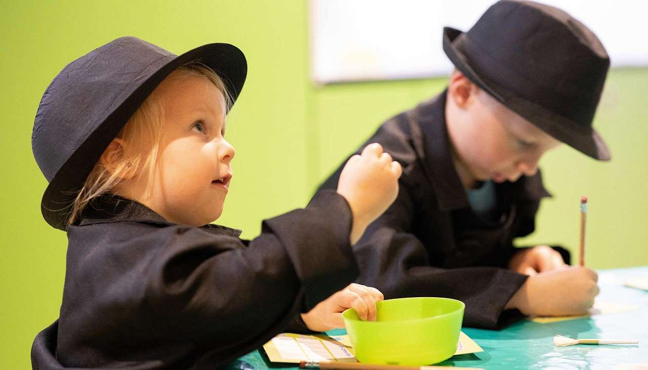 Two children in black hats and coats work at a table; one gestures near a green bowl, the other writes with a pencil. Bright room with lime-green wall and crafts.