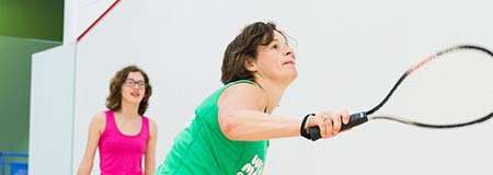 Squash player stretches forward, swinging a racquet to hit the ball; opponent stands behind watching on a bright indoor court with white walls and red boundary lines.