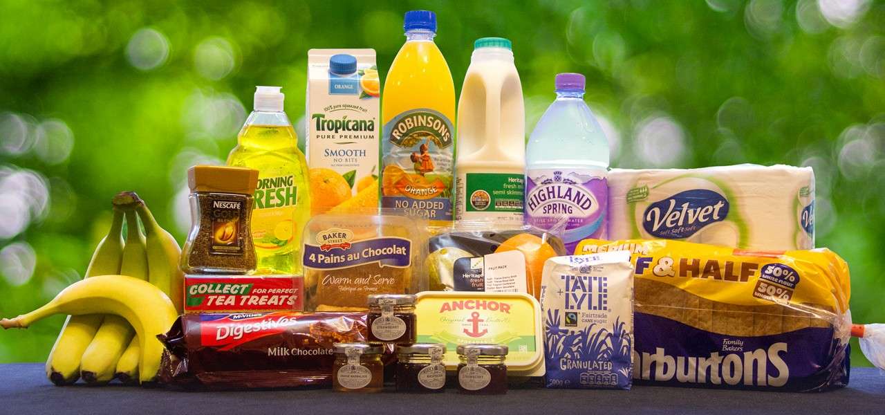 Groceries arranged on a table outdoors. Text visible: NESCAFÉ; Collect Tea Treats; Morning Fresh; McVitie’s Digestives Milk Chocolate; Tropicana Pure Premium Smooth No Bits; Robinsons Orange No Added Sugar; Semi Skimmed; Highland Spring; Velvet; BAKER 4 Pains au Chocolat Warm and Serve; Anchor; Tate & Lyle Fairtrade Granulated; Warburtons Family Bakers Half & Half; 50% white flour 50% wholemeal flour.