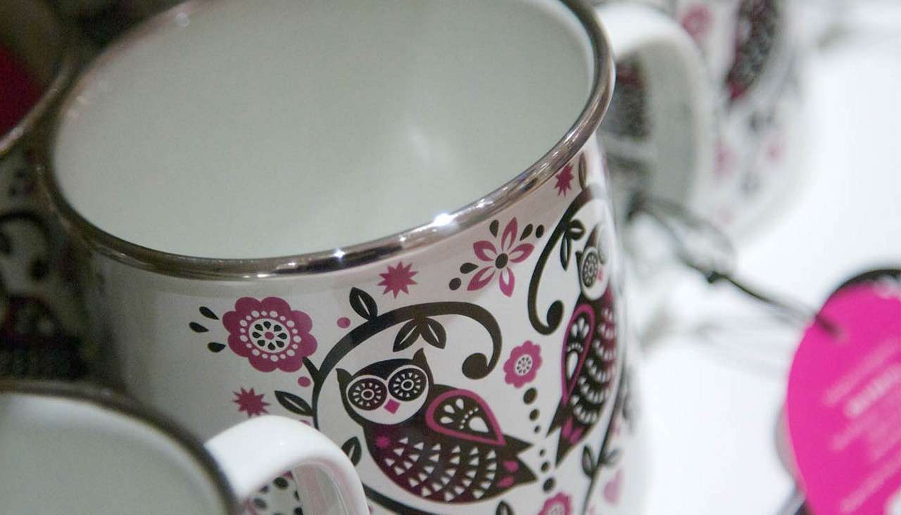 Decorative ceramic mug displays pink-and-black owl and floral patterns; sitting among similar mugs on a shelf, its silver rim gleams in a close-up product display.