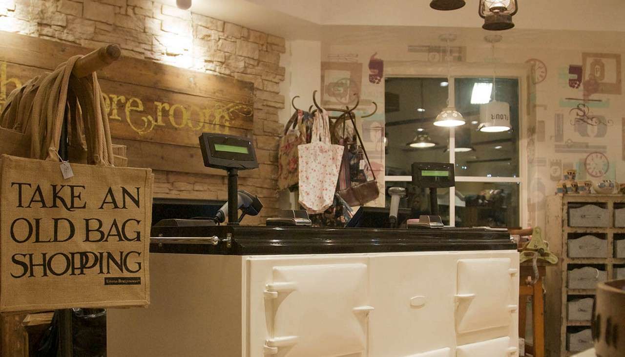 Jute tote hangs beside checkout counter; registers sit on a vintage cream stove. Cozy boutique surrounds with hanging bags, warm lights, and rustic décor. Text: “TAKE AN OLD BAG SHOPPING” “Emma Bridgewater”.
