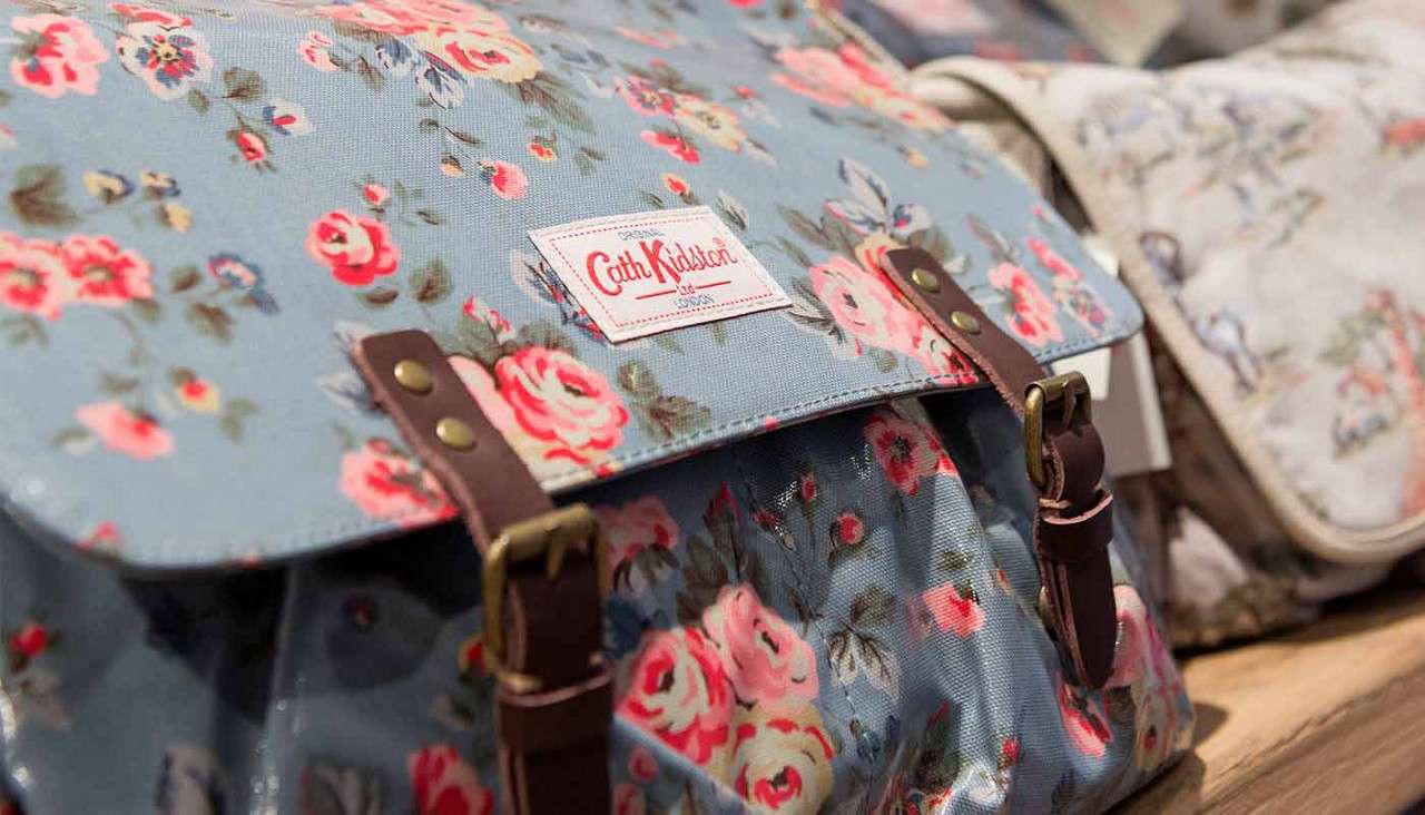 Floral satchel rests on a wooden surface, secured by leather buckle straps, with other patterned bags nearby. Text: "Cath Kidston".